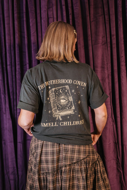 A person shown from the back wearing a dark charcoal t-shirt with a gothic-themed graphic print that reads 'THE MOTHERHOOD COVEN SMELL CHILDREN!' and features an illustration of an eye in a book. The person is standing in front of a dark purple curtain.