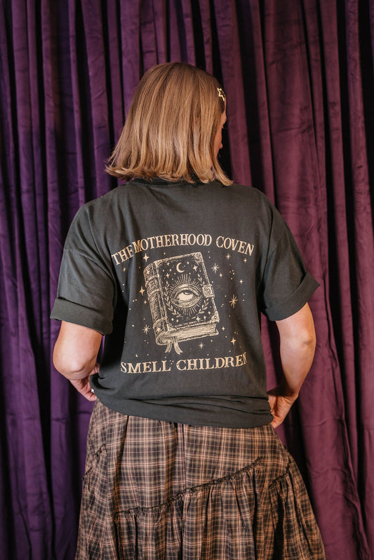 A person shown from the back wearing a dark charcoal t-shirt with a gothic-themed graphic print that reads 'THE MOTHERHOOD COVEN SMELL CHILDREN!' and features an illustration of an eye in a book. The person is standing in front of a dark purple curtain.