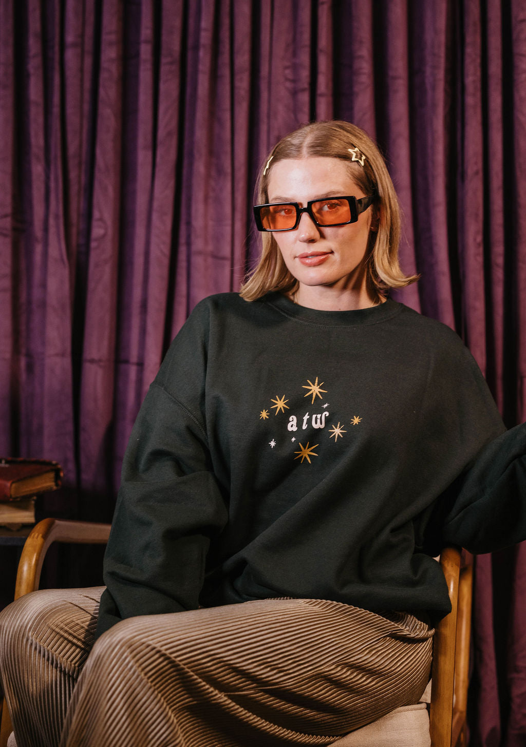 A woman wearing a dark oversized sweatshirt with a gold star and text graphic, paired with brown pinstriped trousers, sitting in a wooden chair against a purple curtain background.
