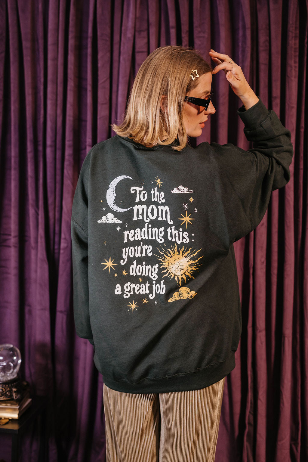 A woman wearing a dark charcoal gray sweatshirt with a celestial graphic and the text 'To the mom reading this: you're doing a great job' in white, set against a dark purple curtain background.
