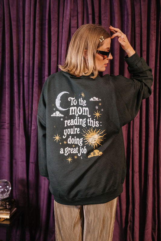 A woman wearing a dark charcoal gray sweatshirt with a celestial graphic and the text 'To the mom reading this: you're doing a great job' in white, set against a dark purple curtain background.
