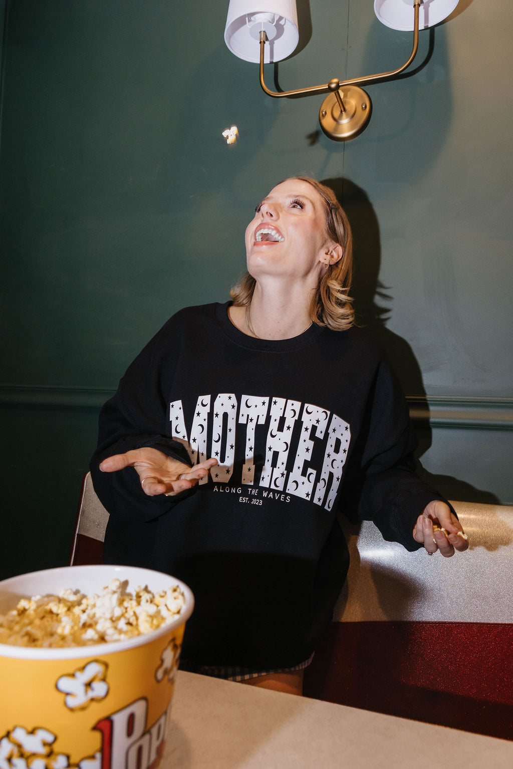 A woman wearing a black crewneck sweatshirt with 'MOTHER' printed in distressed white letters across the chest, looking up toward a wall sconce while holding popcorn in a yellow bucket in the foreground.