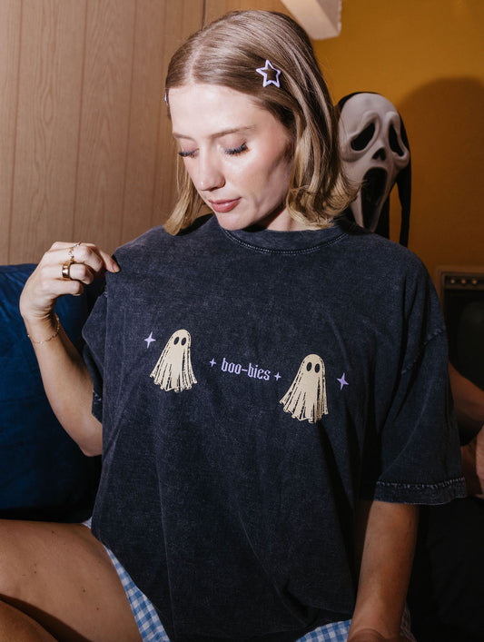 A woman wearing a dark charcoal gray t-shirt with a distressed wash, featuring a graphic of two beige ghosts and the text 'boo-bics' in purple. She is holding the shirt up to show the front print.