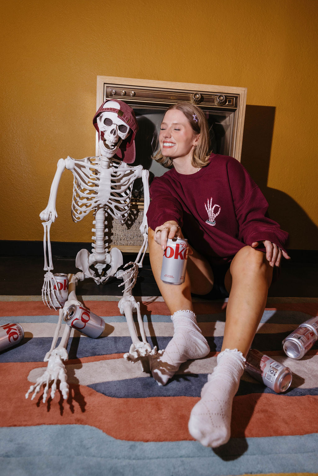 A woman in a maroon sweatshirt and white socks sits on a striped rug next to a life-sized skeleton prop wearing a red cap, with empty soda cans scattered around in a styled indoor setting.