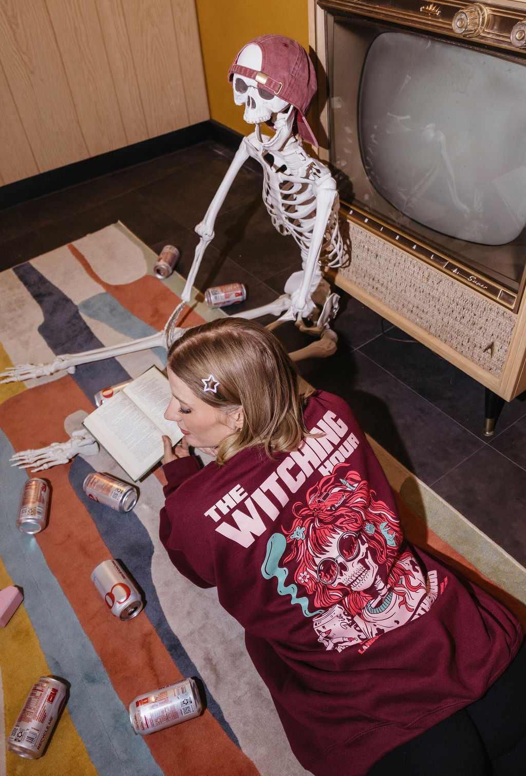 A woman wearing a burgundy graphic t-shirt with 'THE WITCHING HOUR' and a witch illustration lies on a patterned rug next to a skeleton decoration and an old television. Multiple beverage cans are scattered on the floor.