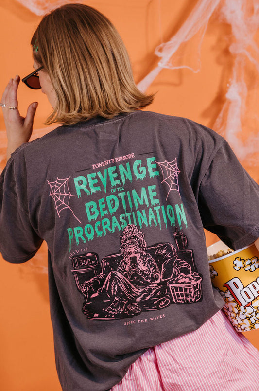 A woman shown from behind wearing a dark charcoal gray graphic t-shirt with green and pink text reading 'REVENGE BEDTIME PROCRASTINATION' and holding a popcorn bucket against an orange background with fake cobwebs.