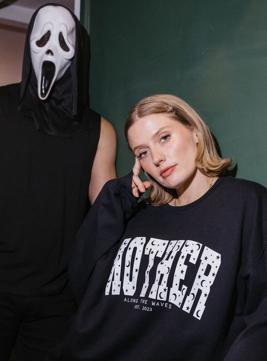 A woman wearing a black crewneck sweatshirt with a white 'MOTHER' graphic stands next to a person in a mask and black hoodie against a dark green wall.
